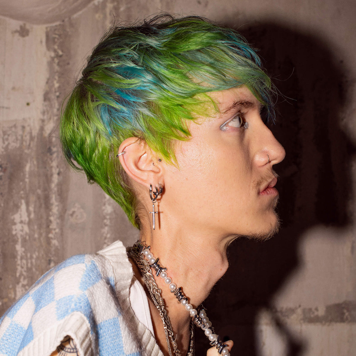 Person mit grün-blauem Haar im Profil, trägt auffällige Ohrringe und Ketten, vor einer rauen Betonwand.