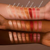 Swatches von NABLA Skin Glazing Highlightern auf drei Hauttönen – acht schimmernde Farben von Champagner bis Kupfer, Rosa und Rot. Namen sind eingeblendet.