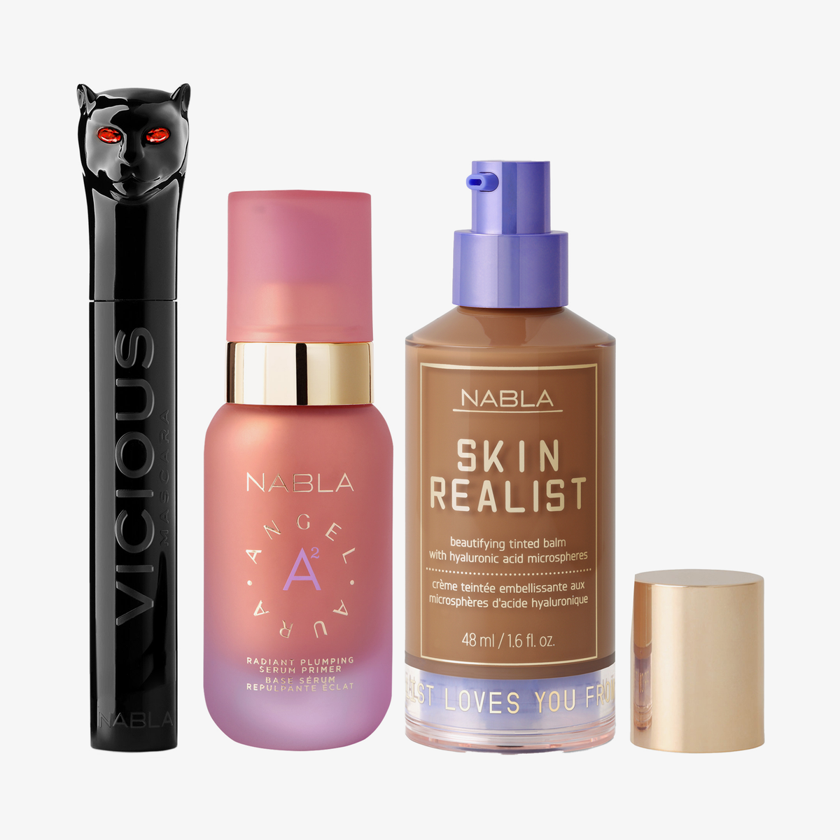 Drei NABLA-Produkte: schwarze Mascara mit Katzenkopf, rosa Angel Aura Serum-Primer und Skin Realist Tinted Balm in der Farbe 6 Dark mit lilafarbenem Pumpspender.