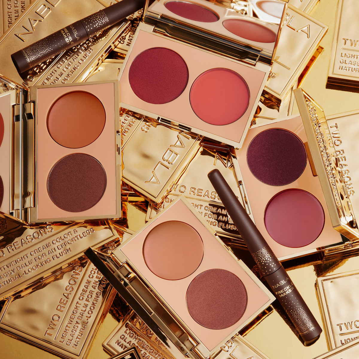 Mehrere geöffnete NABLA „Two Reasons“ Blush-Kompaktpaletten in verschiedenen Farbkombinationen mit pudrigen und cremigen Texturen, dekorativ arrangiert auf glänzenden goldenen Verpackungen. Zwei NABLA Freckle Maker Stifte liegen dazwischen.