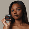 Eine Frau hält den geöffneten Skin Mate Compact von Mesauda Beauty in der Nähe ihres Gesichts und blickt in Kamera.