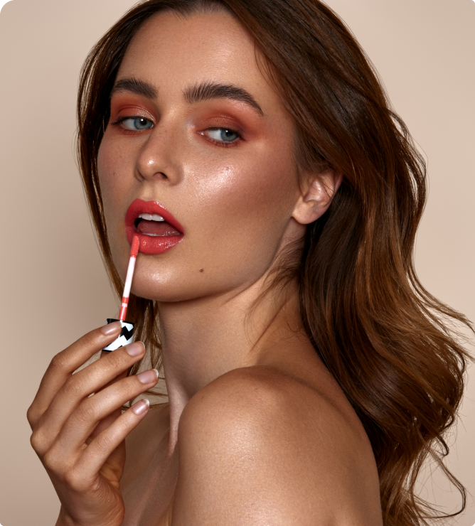 Model mit bronzefarbenem Augen-Make-up trägt glänzenden Lippenstift mit Applikator auf, vor neutralem Hintergrund.