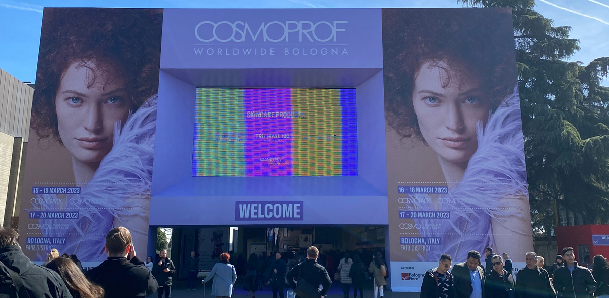 Eingang zur COSMOPROF Messe 2023 in Bologna mit großem Banner, LED-Wand und Besucher:innen vor dem Veranstaltungsort.