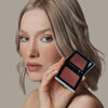 Eine Frau hält den geöffneten Skin Mate Compact von Mesauda Beauty in der Nähe ihres Gesichts und blickt in Kamera.