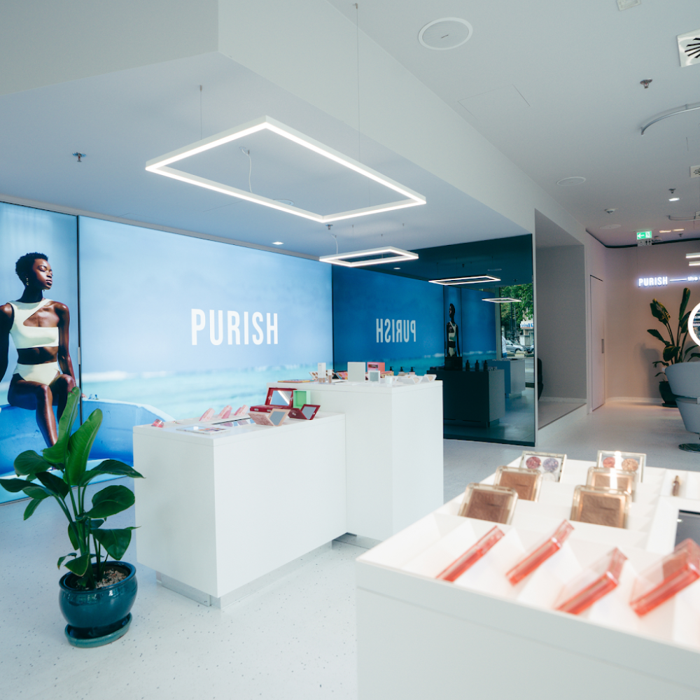 Moderner PURISH Store mit hellen Möbeln, Kosmetikprodukten und großem Wandbild einer Frau vor Meereskulisse.