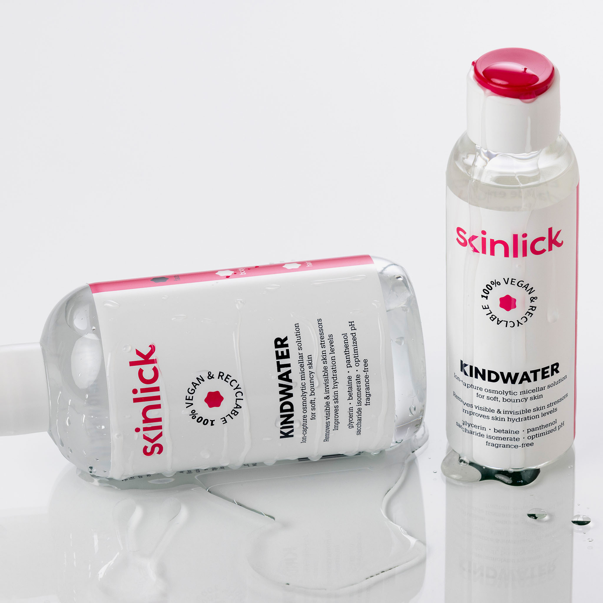 Zwei Flaschen mit skinlick Kindwater, eine stehend und eine liegend, stehen auf einer weißen Fläche mit Wassertropfen.