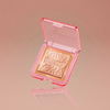 Ein goldener Highlighter in einem rosafarbenen transparenten Kompaktetui mit der Aufschrift NABLA, präsentiert auf einem pfirsichfarbenen Hintergrund.