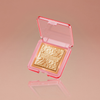 Der NABLA Skin Glazing Highlighter in Gold wird in einem durchsichtigen, rosafarbenen Kompaktetui vor einem zartrosa Hintergrund präsentiert.