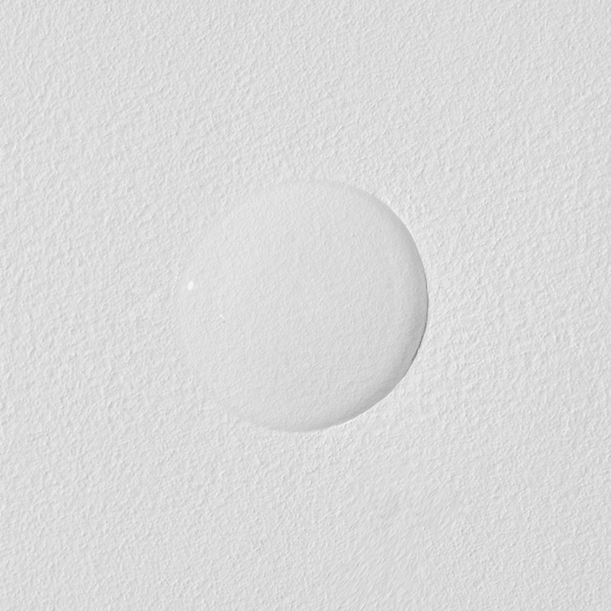 Ein einzelner milchiger Tropfen Advanced Snail 96 Mucin Power Essence auf einer strukturierten weißen Oberfläche.