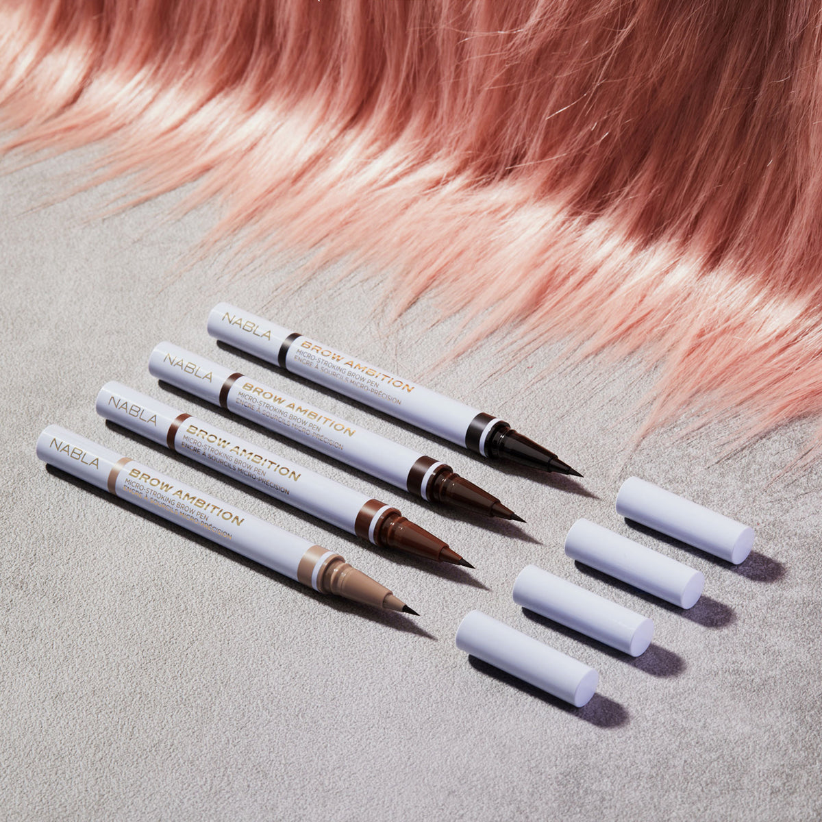 Vier geöffnete Brow Ambition Pens von Nabla Cosmetics in verschiedenen Brauntönen liegen nebeneinander auf grauem Untergrund mit rosafarbenem Kunsthaar im Hintergrund, daneben die abgenommenen Verschlusskappen.
