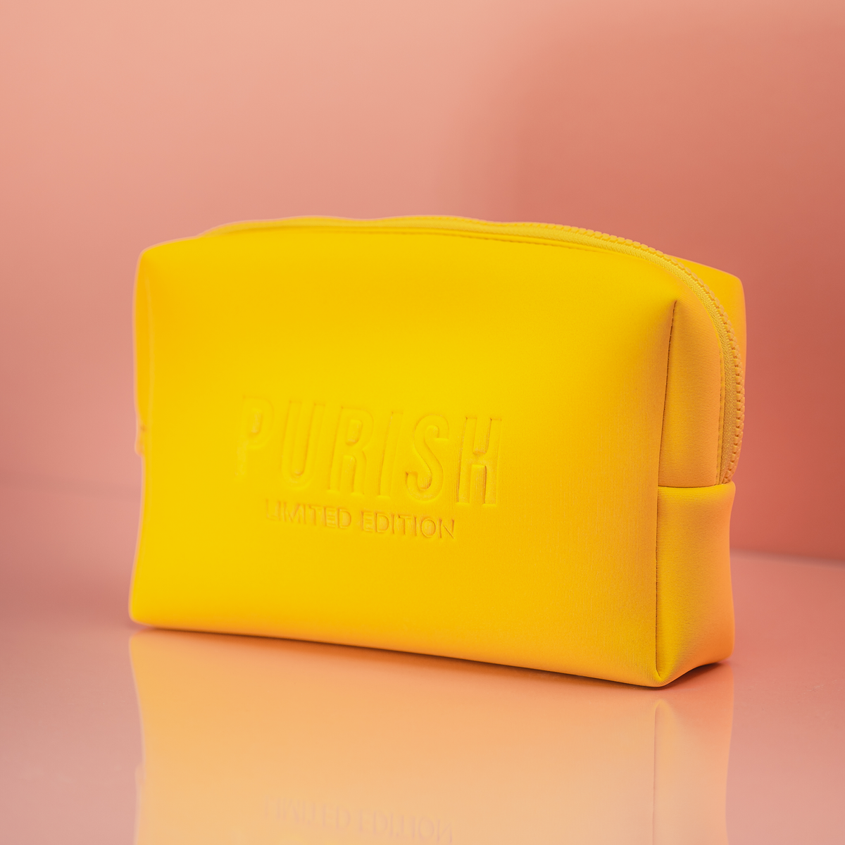 Die leuchtend gelbe PURISH Bag mit geprägtem Markenzeichen, wird vor einem rosa Hintergrund präsentiert.