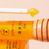 Applikatorstab mit gelbem Lippenbalsam neben einer Tube NUXE Honey Lip Care Rêve de Miel und einem hölzernen Honiglöffel.