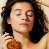 Eine Frau mit lockigem Haar hält eine Flasche NUXE Huile Prodigieuse® Florale, schließt die Augen und sieht ruhig und entspannt aus.