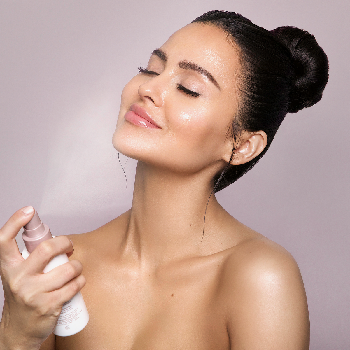 Lächelnde Frau mit glatter Haut und dunklem Haar im Dutt sprüht Ultra Hydrating Fine Mist auf ihr Gesicht, die Augen geschlossen.