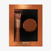 Danessa Myricks Beauty Yummy To Go Kit mit Yummy Skin Glow Serum und Blurring Balm Powder in einer kupferfarbenen Verpackung.