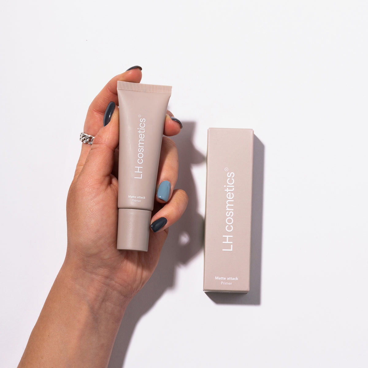 Eine Hand hält eine Tube Matte Attack Primer von LH Cosmetics neben der passenden Schachtel auf weißem Hintergrund.