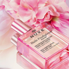 Die Flasche NUXE Huile Prodigieuse® Florale hat einen rosafarbenen Verschluss und ist von zartrosa Blumen umgeben.