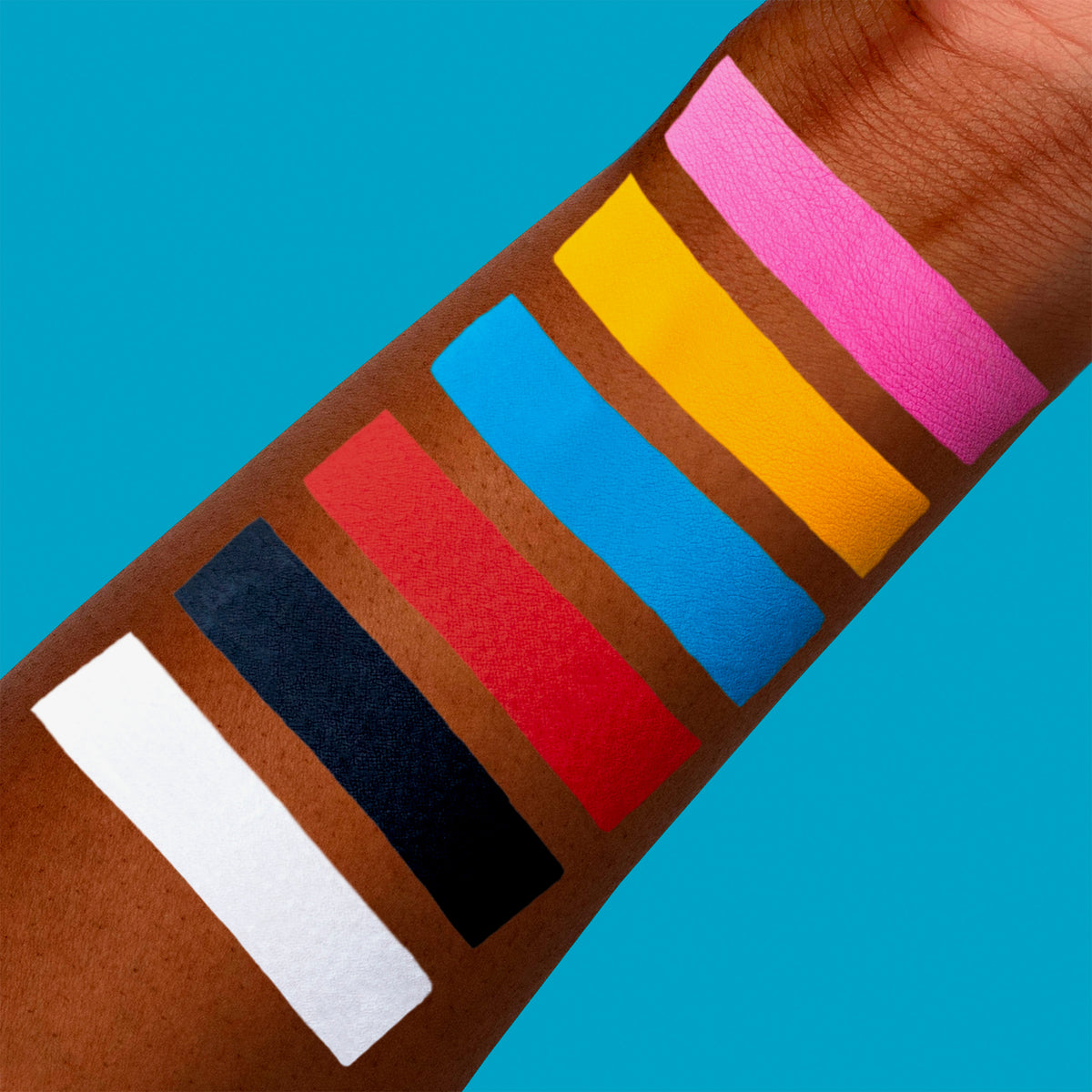 Das Bild zeigt einen Arm mit sieben deckenden Farbswatches der SUVA Beauty OPAKES Cosmetic Paints; aufgetragen auf dunkler Haut vor blauem Hintergrund.