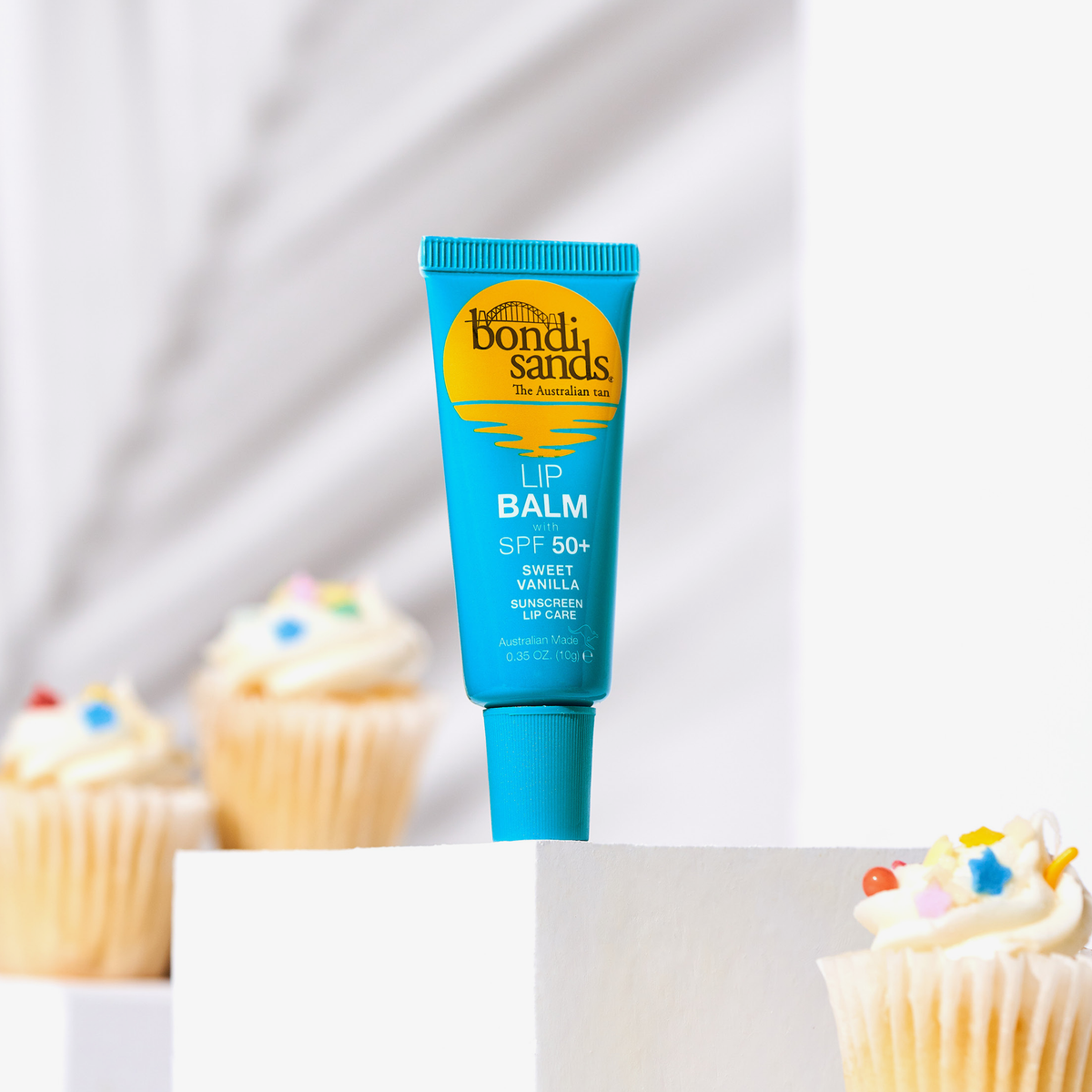 Eine Tube Lip Balm SPF 50+ Sweet Vanilla in Blau steht auf einer weißen Oberfläche neben Cupcakes mit Streuseln.