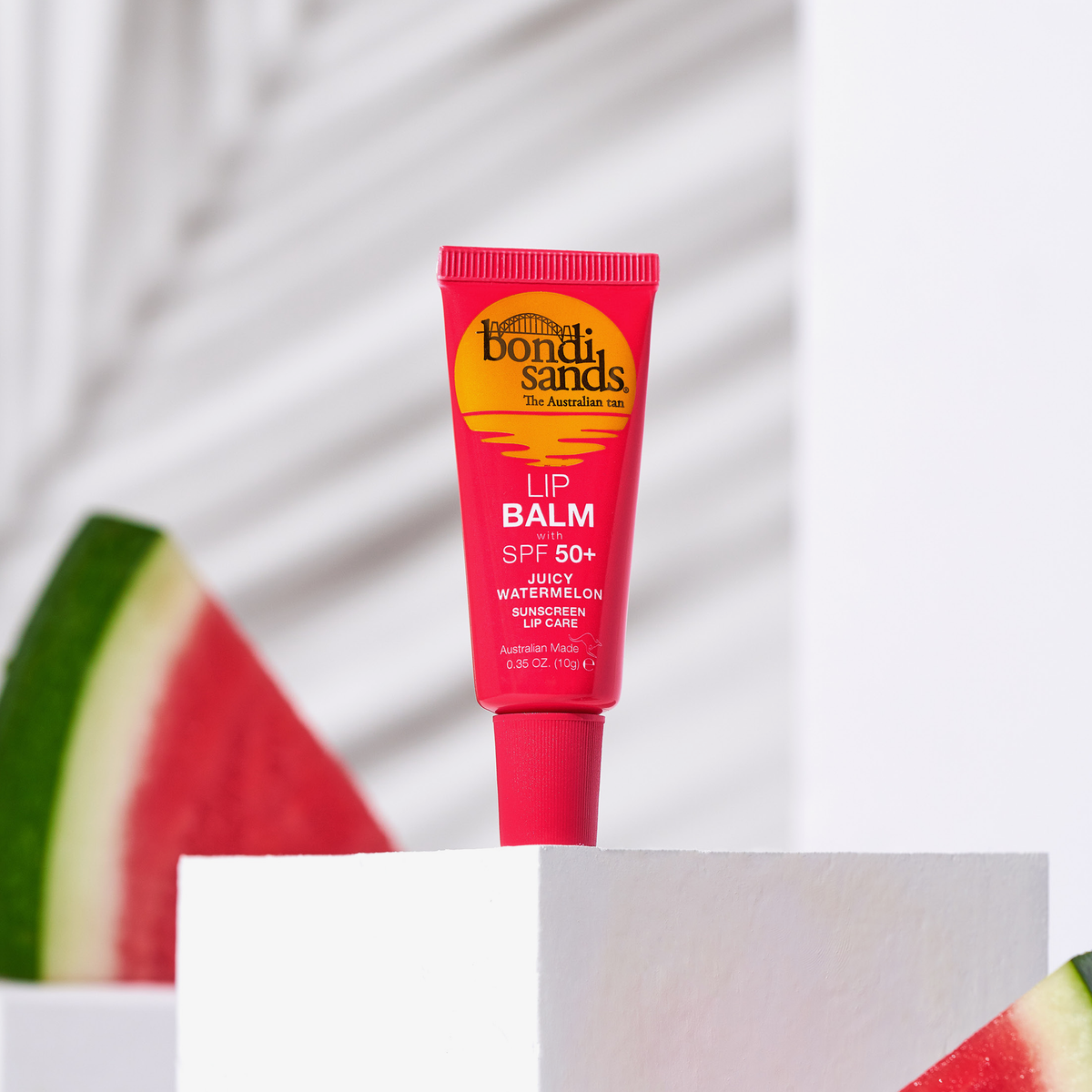Eine rote Tube Lip Balm SPF 50+ Juicy Watermelon von Bondi Sands steht auf einem weißen Block, umgeben von Wassermelonenscheiben