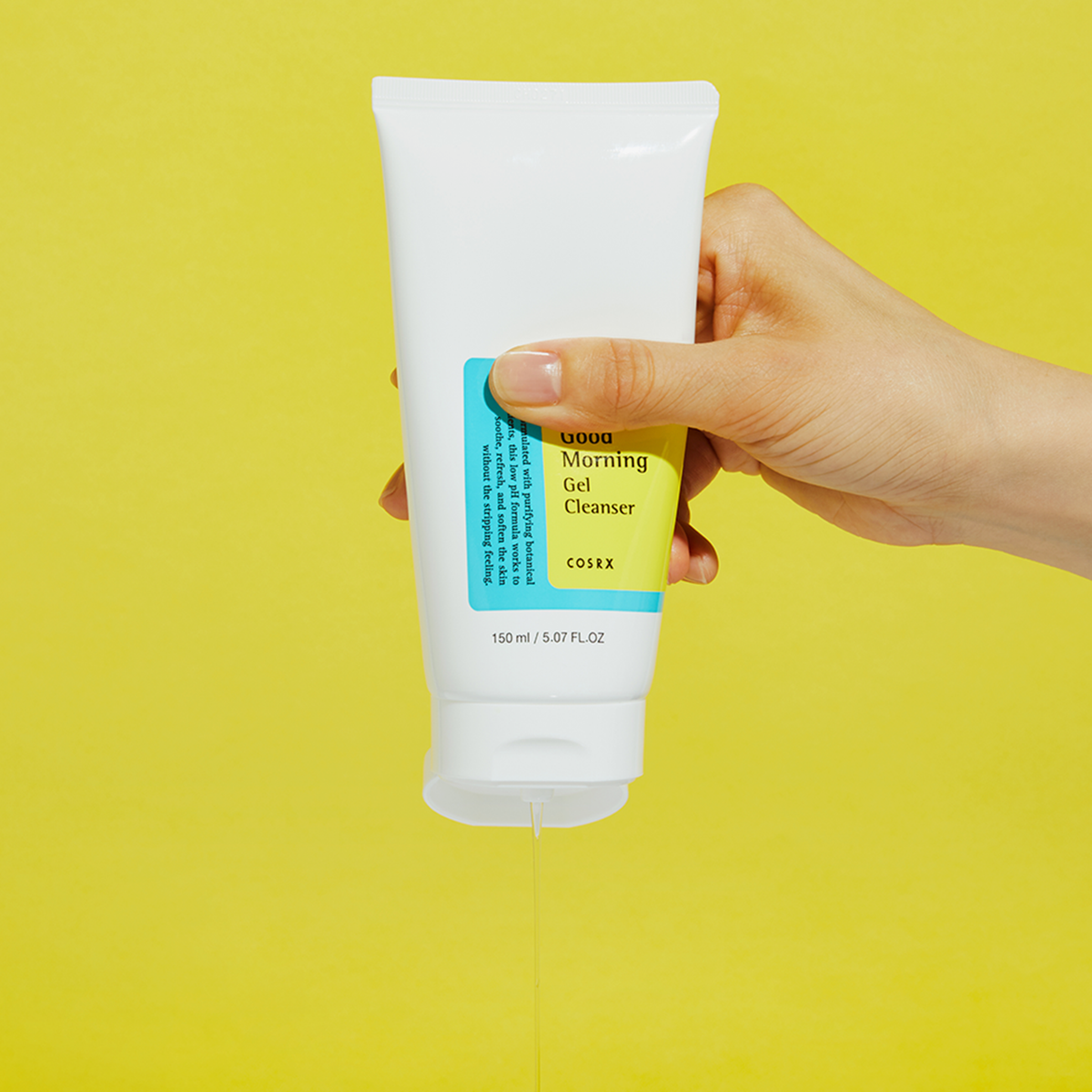 Eine Hand drückt Low-pH Good Morning Gel Cleanser aus einer weißen Tube vor einem leuchtend gelben Hintergrund.