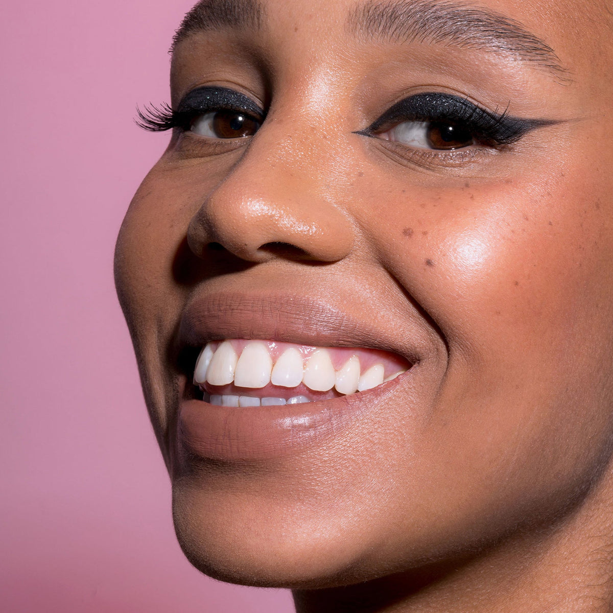 Eine Frau lächelt mit geflügeltem Eyeliner und natürlichem Make-up. Sie trägt den Crayon Lipliner von LH Cosmetics vor einem rosa Hintergrund.