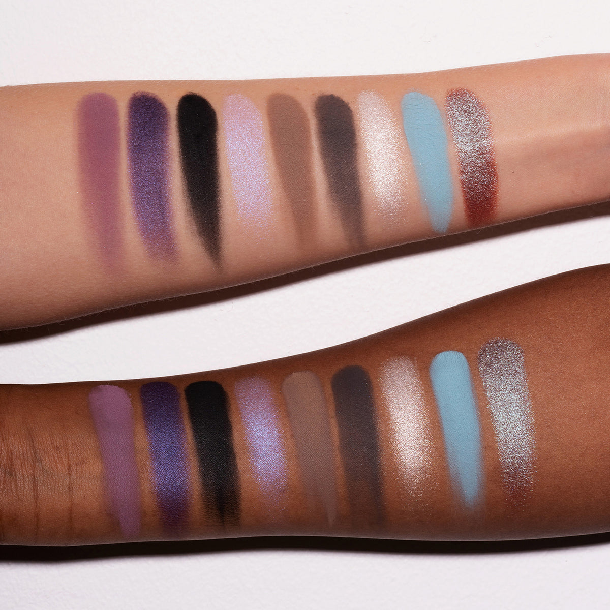 Zwei Arme mit unterschiedlichen Hauttönen zeigen lilane, schwarze, grau und beige Farbmuster der Daybreak Palette von LH Cosmetics.