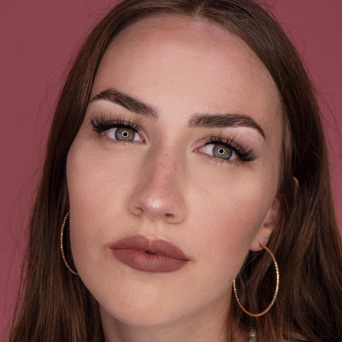 Model mit langen braunen Haaren, Creolen und neutralem Make-up trägt NICLAY HD Soft Lashes vor einem rosa Hintergrund.