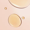 Nahaufnahme der beigen Tröpfchen der Honey Lip Care Rêve de Miel von NUXE auf einer glatten, blassrosa Oberfläche.