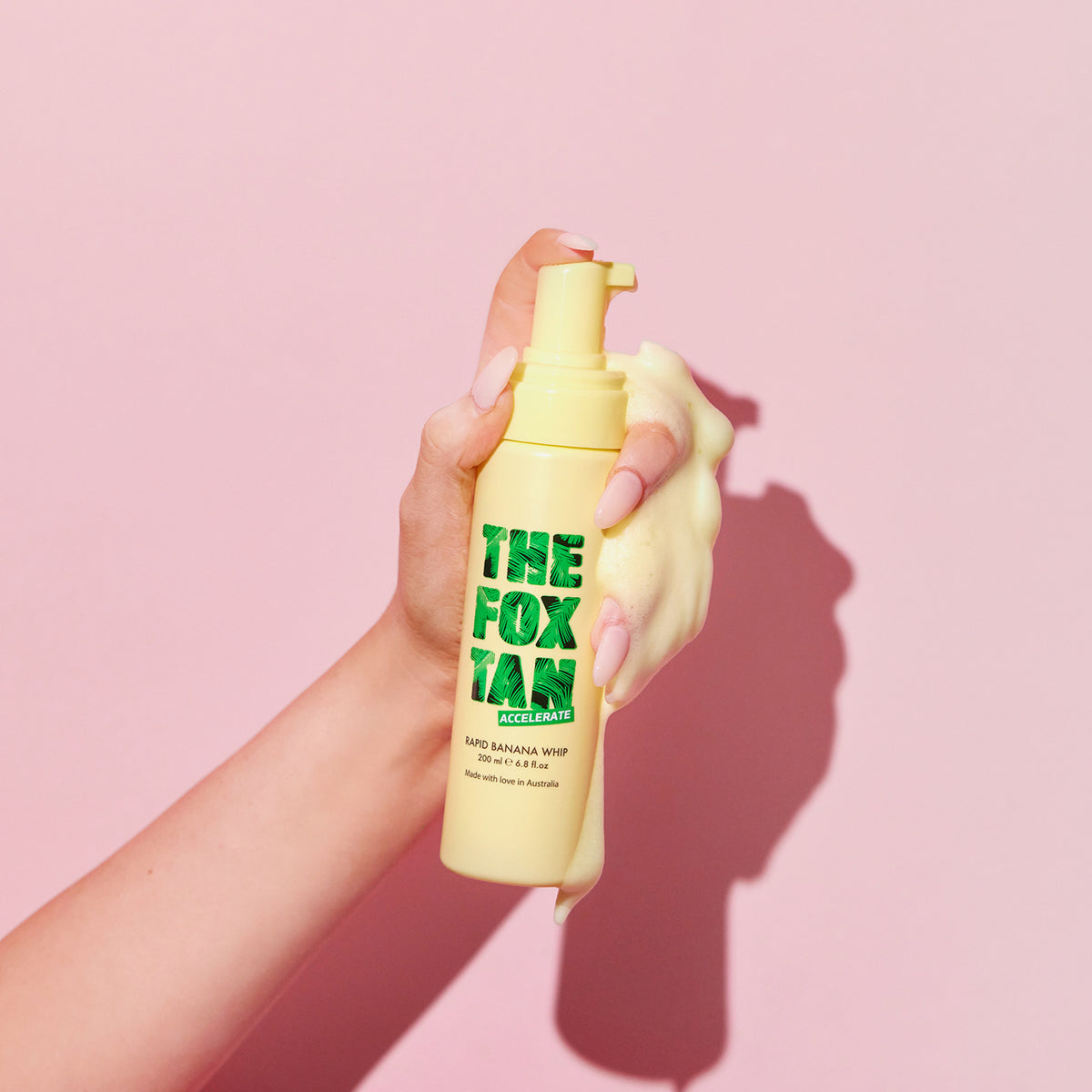 Eine Hand hält die gelbe Rapid Banana Whip-Flasche vor einem rosa Hintergrund.