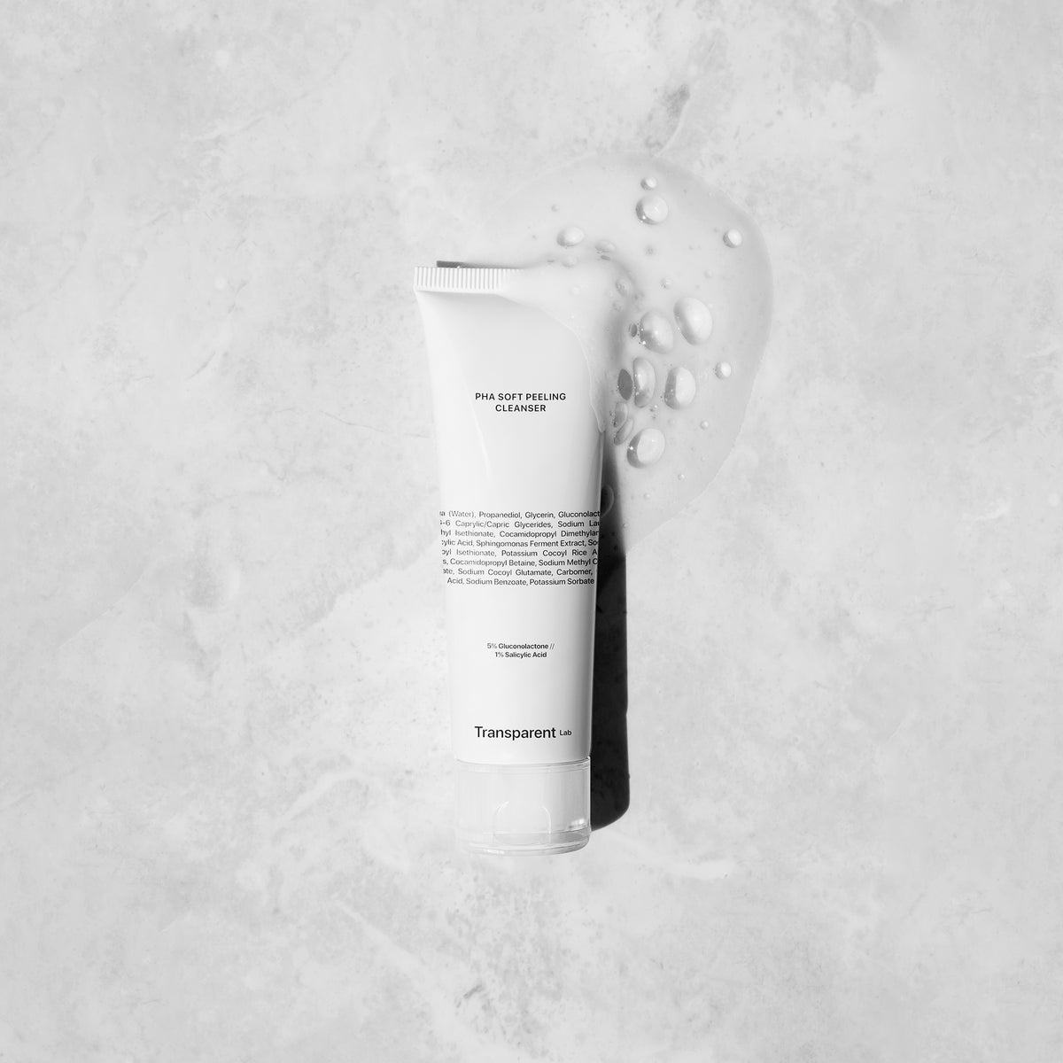 Der PHA Soft Peeling Cleanser von Transparent liegt auf einer grauen Oberfläche, daneben ist Flüssigkeit verschüttet.