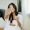 Frau in weißem Outfit sitzt auf einem Sofa und hält lächelnd den Wide Awake Full Coverage Concealer von Dominique mit rosafarbener Kappe in die Kamera.