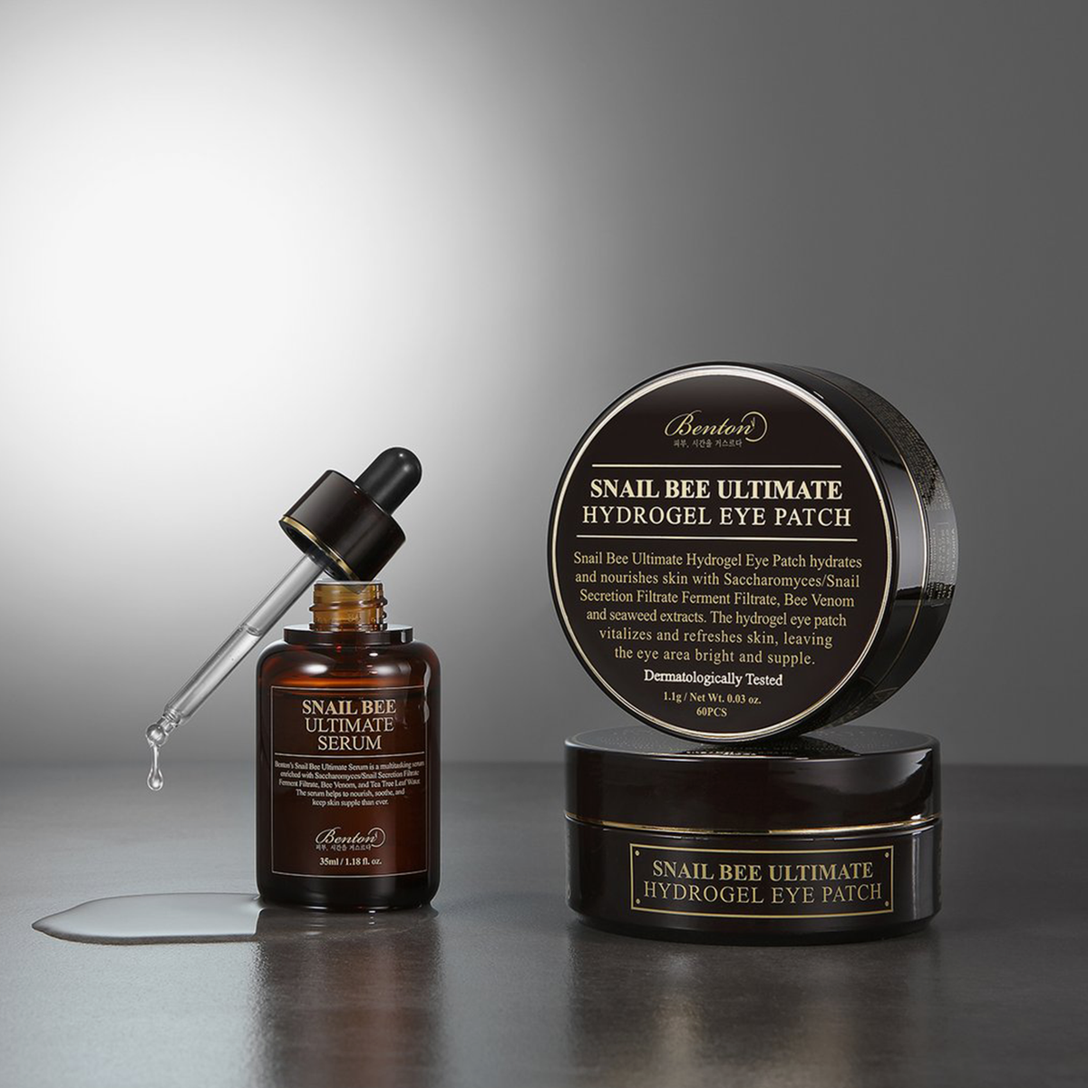 Flasche Snail Bee Ultimate Serum mit offenem Pipettenapplikator, neben 2 Snail Bee Eyepatch Tiegeln in grauer Umgebung