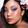 Frau trägt lila Make-up-Look aus der Daybreak Palette von LH Cosmetics und goldene Creolen, mit glänzender Haut und Blumen im Haar.