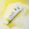 Eine Tube Pure Tøday AHA/PHA Peeling Cleanser auf einem schaumigen gelben Flüssigkeitshintergrund.