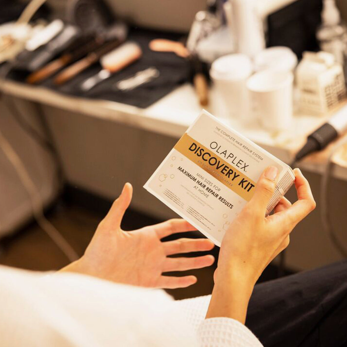 Eine Person hält das Olaplex Discovery Kit in einem Salon, im Hintergrund sind auf der Theke verschiedene Haarwerkzeuge und -produkte ausgestellt.