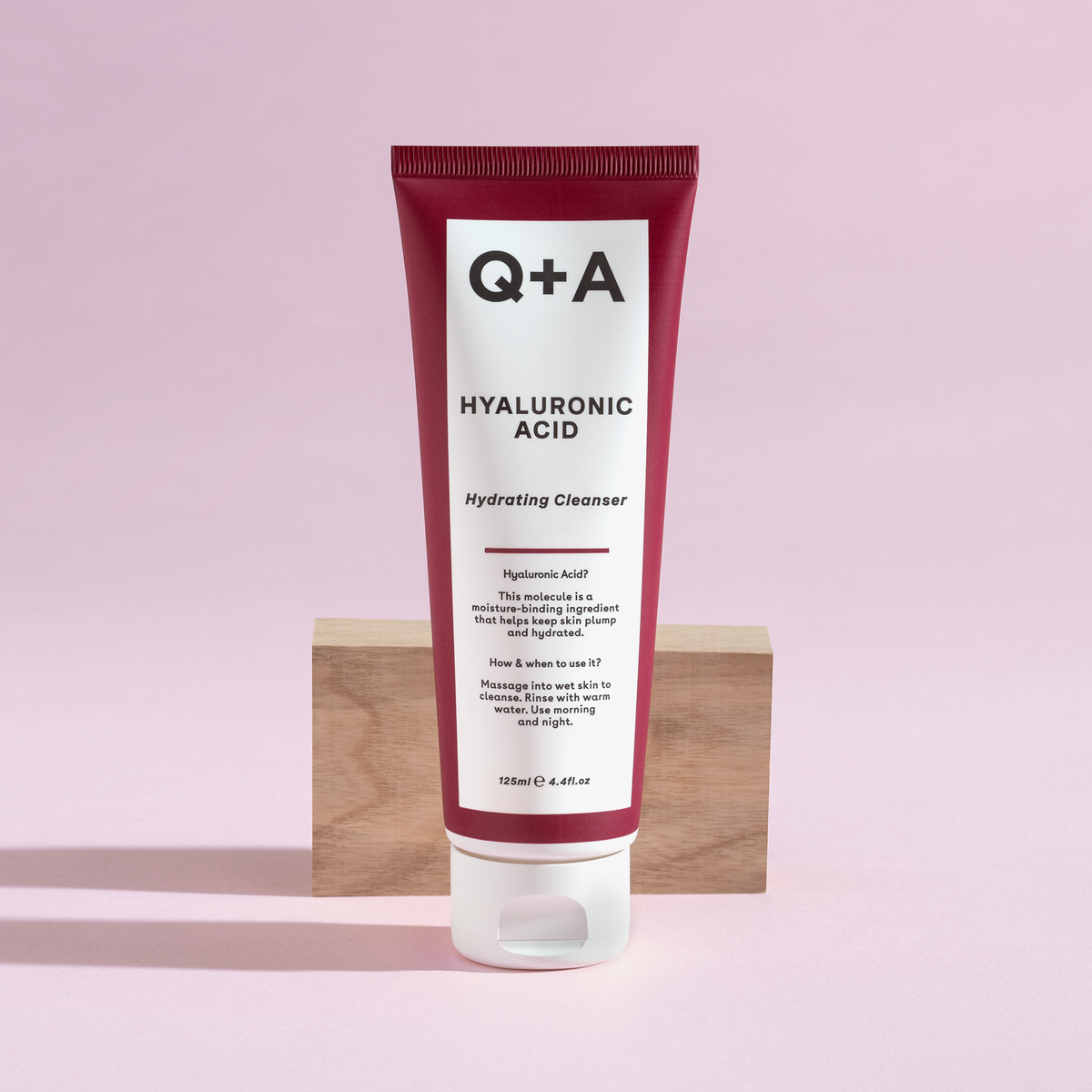 Eine Tube Hyaluronic Acid Hydrating Cleanser in Rot und Weiß steht aufrecht vor einem Holzblock auf rosa Hintegrund.