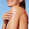 Eine Frau in einem roten Badeanzug lächelt, während sie unter einem klaren blauen Himmel die NUXE Sun Delicious Lotion auf ihre Schulter aufträgt.