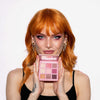 Eine Frau mit orangefarbenem Haar hält die geöffnete Blomma Palette von LH Cosmetics, trägt dazu passendes rosa Make-up und ein schwarzes Oberteil.