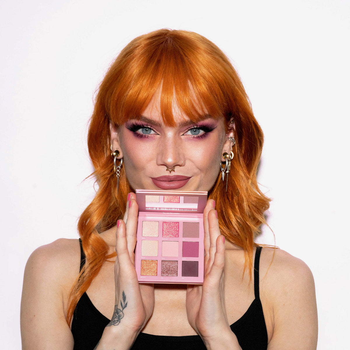 Eine Frau mit orangefarbenem Haar hält die geöffnete Blomma Palette von LH Cosmetics, trägt dazu passendes rosa Make-up und ein schwarzes Oberteil.