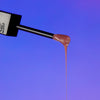 Der Applikator des Gloss Matrix Lipgloss von Mesauda Beauty mit einer tropfenden Spitze vor blauem Hintergrund.