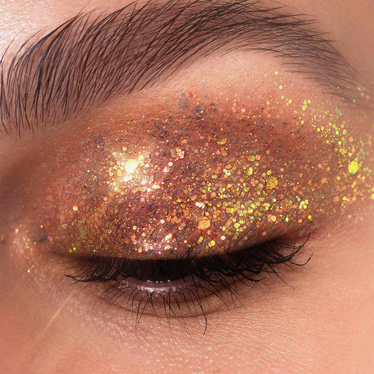 Geschlossenes Auge geschmückt mit dickem, glitzerndem und goldenem Lidschatten aus der Glorious Lights Glitter Palette, mit langen, dunklen Wimpern.