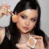 Eine Frau trägt mit einem Make-up-Pinsel OFRA Cosmetics Pure Glow Finishing Powder auf ihre Wange auf und hält das Kompaktpuder in der Hand.