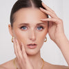 Eine Frau mit zurückgekämmtem Haar, Goldschmuck und natürlichem Make-up, das mit der Essential Palette kreiert wurde, posiert mit ihren Händen, die ihr Gesicht umrahmen.