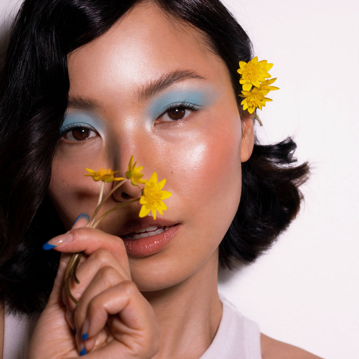 Frau mit blauem Lidschatten aus der Daybreak Palette von LH Cosmetics hält gelbe Blumen vor ihr Gesicht, eine weitere Blume steckt in ihrem dunklen, welligen Haar.