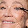 Eine ältere Frau lächelt, während sie LH Cosmetics Infinity Power Lash Lengthening Mascara auf ihre oberen Wimpern aufträgt.