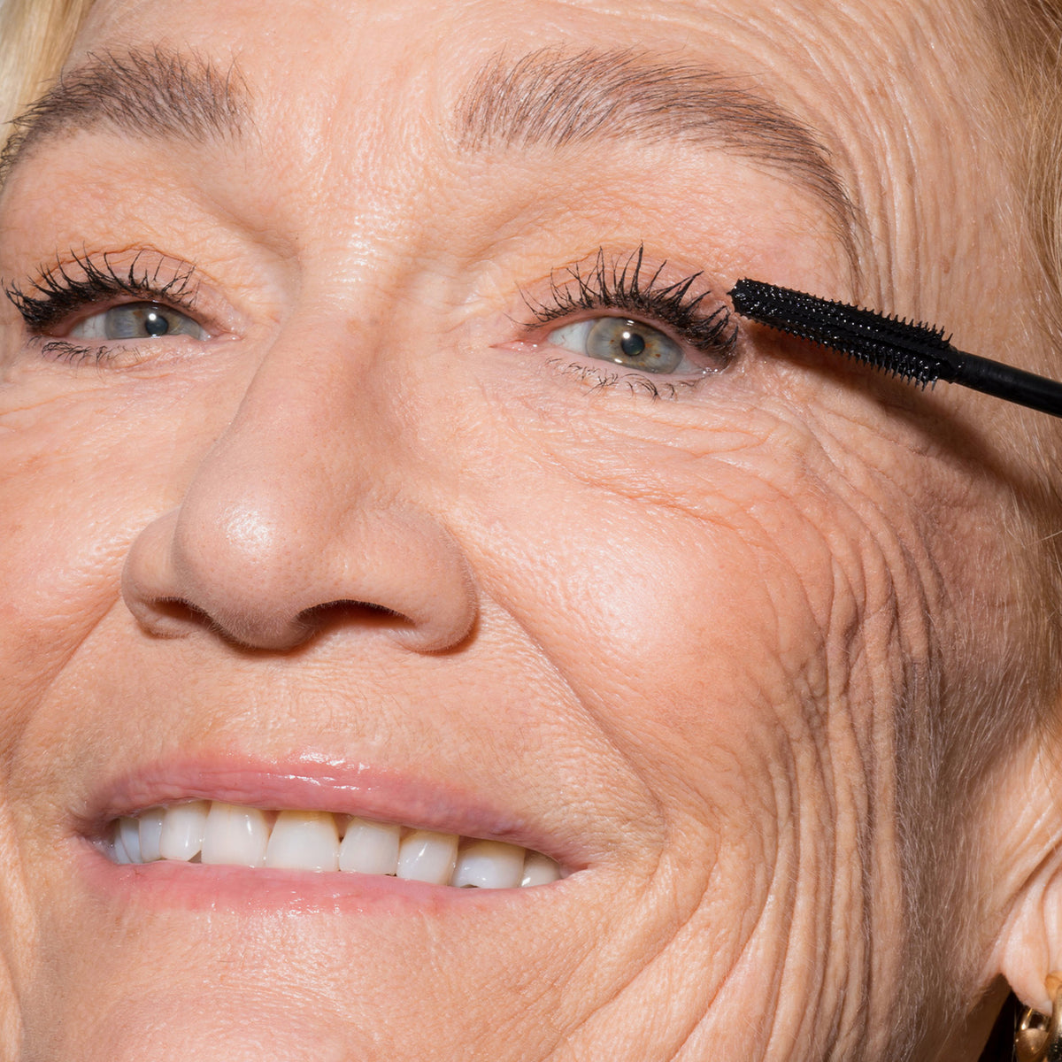 Eine ältere Frau lächelt, während sie LH Cosmetics Infinity Power Lash Lengthening Mascara auf ihre oberen Wimpern aufträgt.