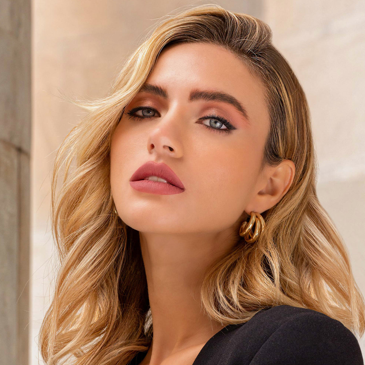 Eine Frau mit gewelltem blondem Haar, goldenen Ohrringen und natürlichem Make-up trägt Mesauda Beauty Cult Matte Lipstick und blickt in die Kamera.