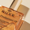 Nahaufnahme einer Flasche NUXE Huile Prodigieuse® OR, deren golden schimmerndes Öl im Sonnenlicht glitzert.