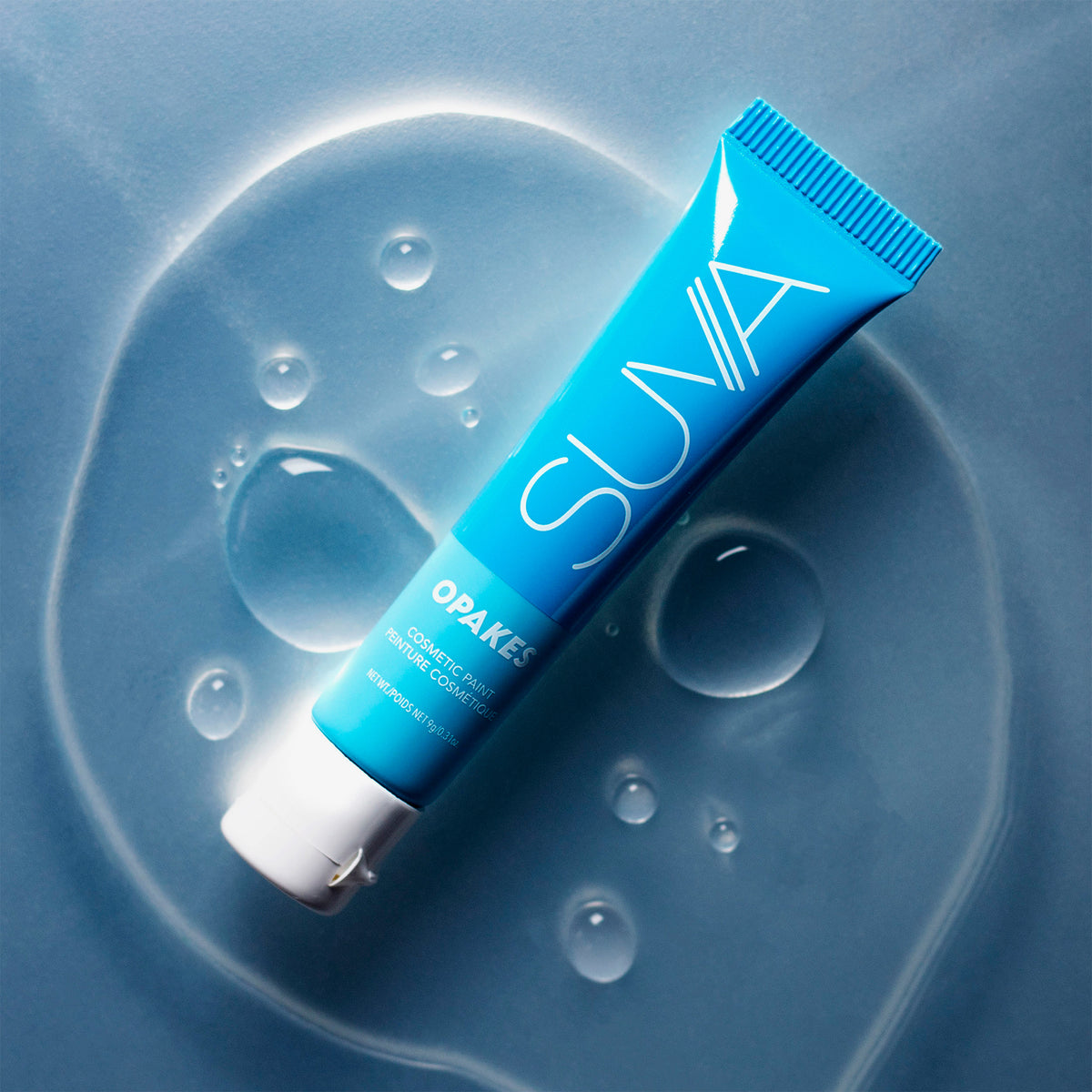 Eine blaue Tube mit der Aufschrift „Opakes Cosmetic Paint“ liegt auf einer nassen blauen Oberfläche mit Wassertropfen.